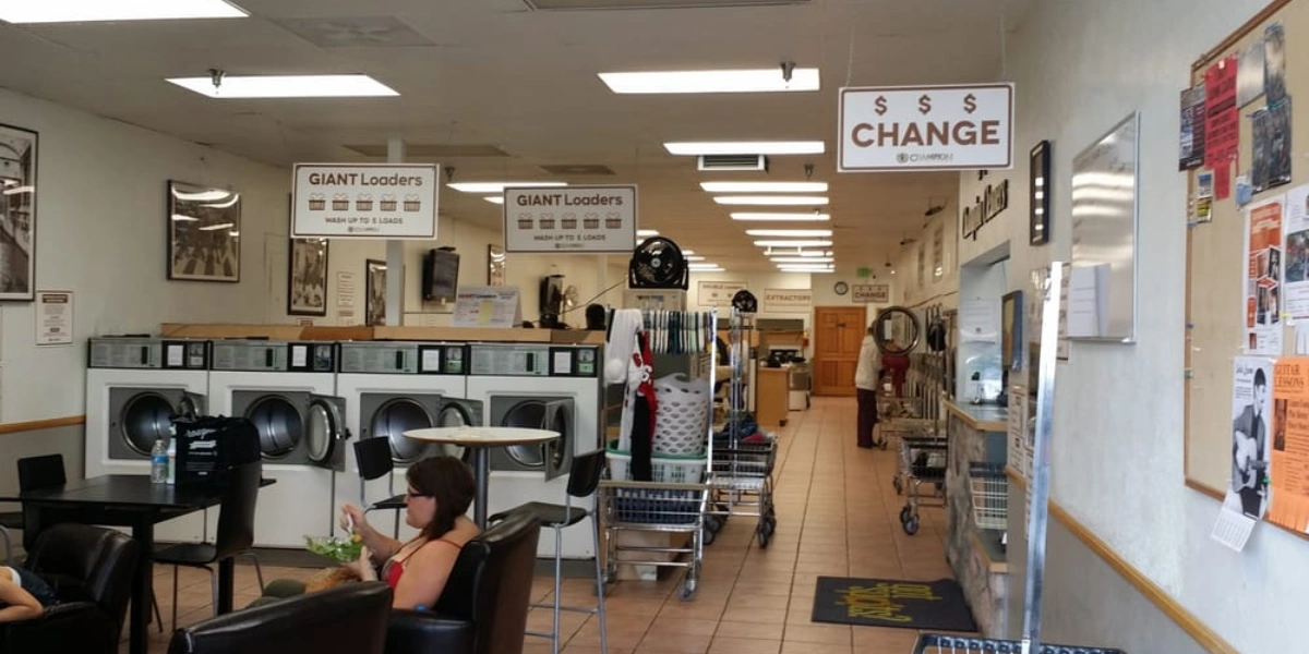 Bright laundromat interior with rows of industrial washers and dryers, a change machine, laundry carts, and a customer seating area.